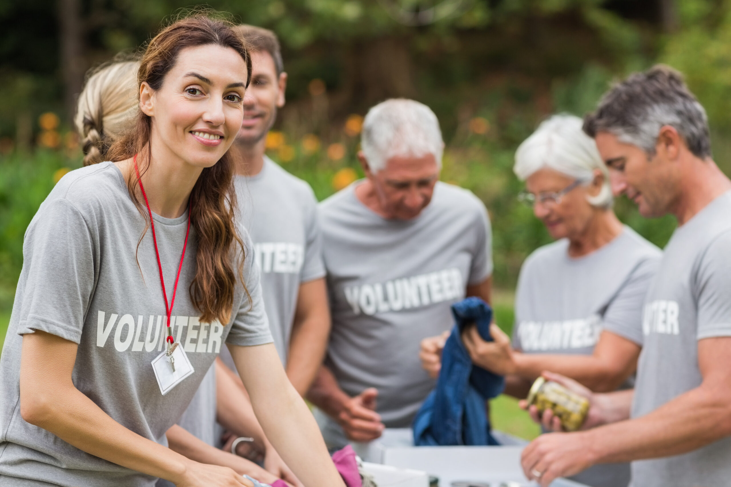 A smiling woman in a "VOLUNTEER" t-shirt leads a group of volunteers in an outdoor community service project.