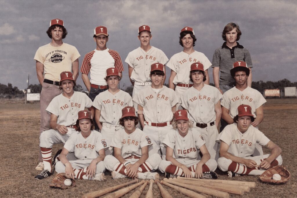 Vintage youth baseball team named Tigers posing on a field with bats and gloves arranged in front; players wear white uniforms with red caps and lettering, coaches stand behind under a cloudy sky.