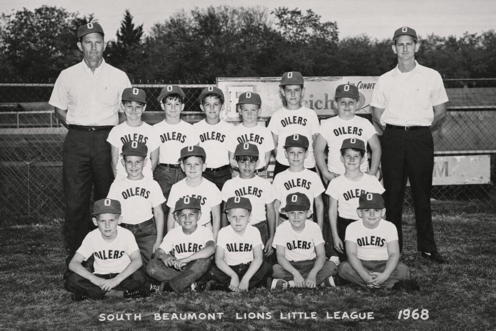 Black‑and‑white photograph of the 1968 South Beaumont Lions Little League “Oilers” Championship team, showing thirteen boys and two adult coaches standing and kneeling in front of a chain‑link fence. The assistant coach on the far right is John’s father.