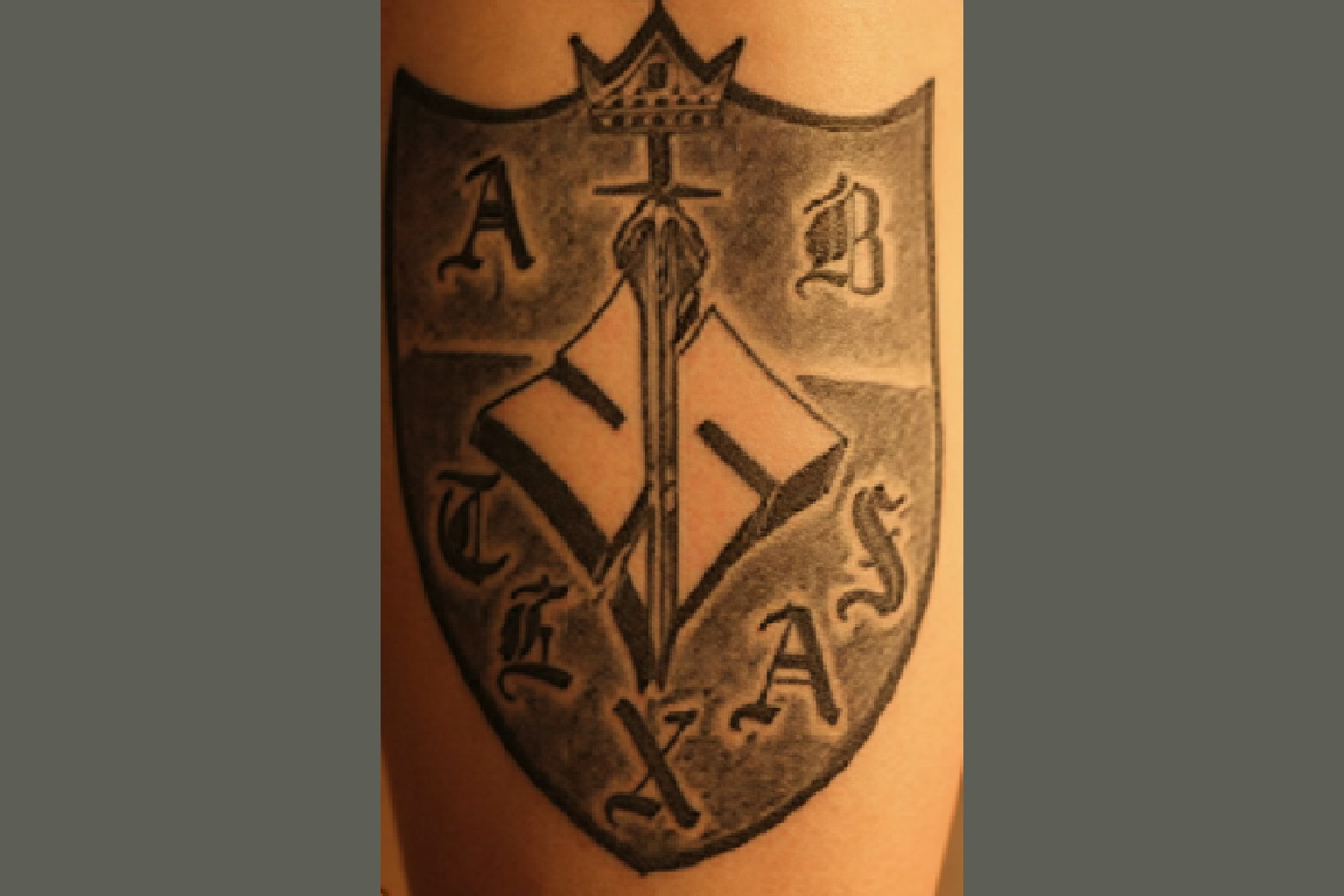 A black ink tattoo on skin depicting a shield-shaped emblem of the Aryan Brotherhood of Texas. Inside the shield is a central broadsword pointing downwards, overlaid on a large, stylized geometric swastika symbol. The letters 